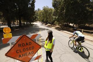 LOS ANGELES, CA - JUNE 29, 2022 - - A bicyclist rides past city worker Emma Delgado to head down a one-third mile portion of Griffith Park Drive that is indefinitely closed to car traffic to improve safety for pedestrians and cyclists in Griffith Park in Los Angeles on June 29, 2022. Delgado was on site to tell motorists that they could not go down this portion of Griffith Park Drive. The road is known to draw high-speed drivers who are looking for a shortcut, avoiding the 134 and 5 freeways. The temporary closure has been in place since Monday, June 27 and will carry on indefinitely until more permanent changes to the park's infrastructure are in place. (Genaro Molina / Los Angeles Times)