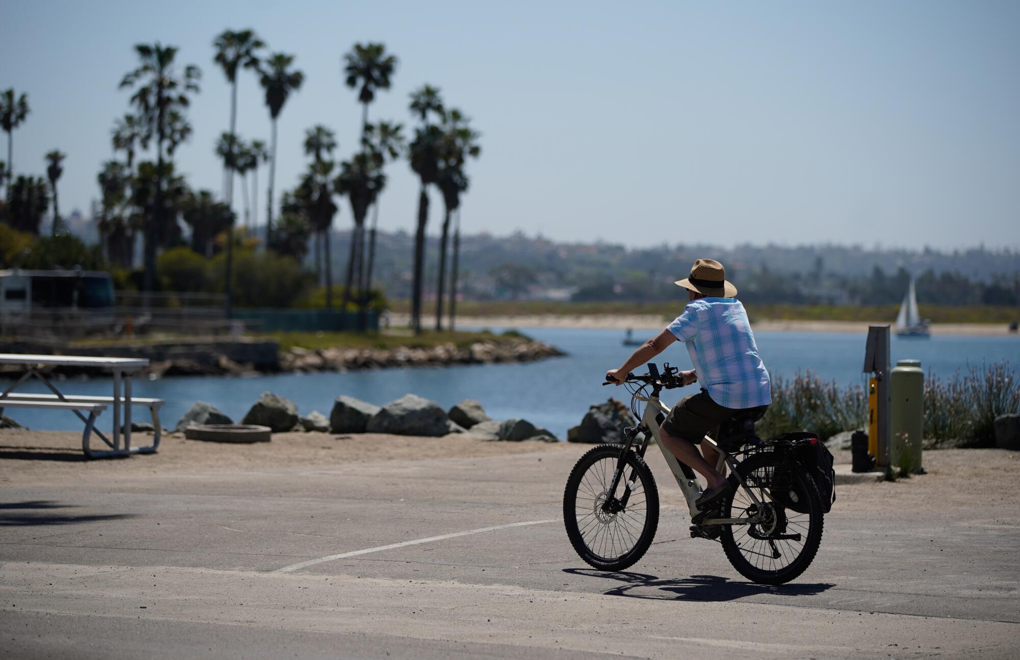 A man in a brimmed hat rides a bicycle on a road along the bay, palm trees and a sailboat in the distance.