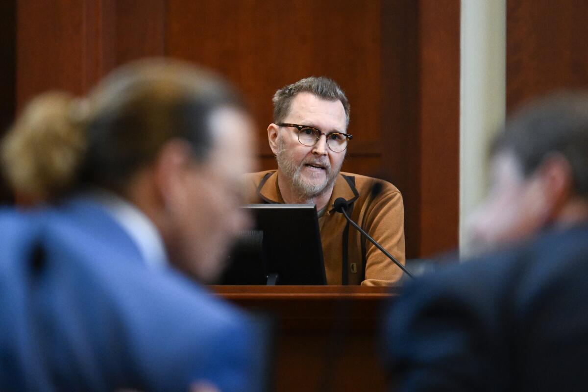 A man in glasses sits on the witness stand behind blurry images of two men conversing in the foreground