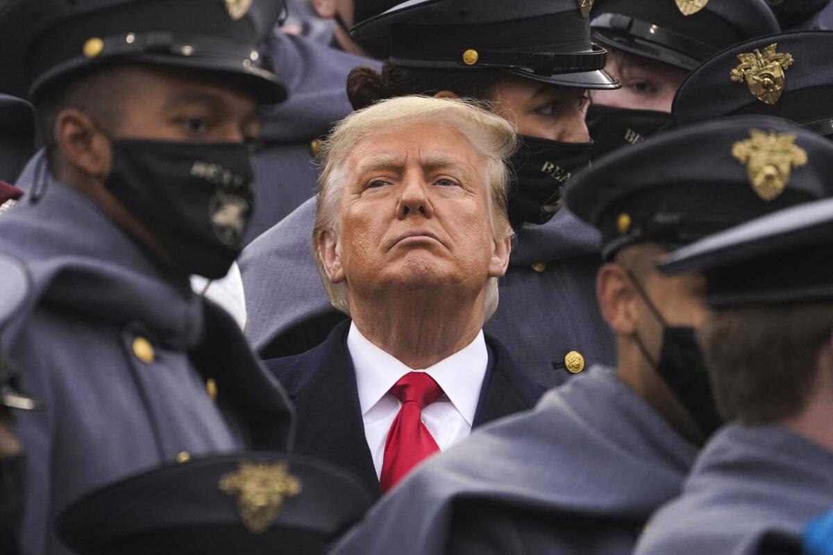 An unmasked man in suit and red tie is surrounded by masked people in dark military uniforms and caps with insignia