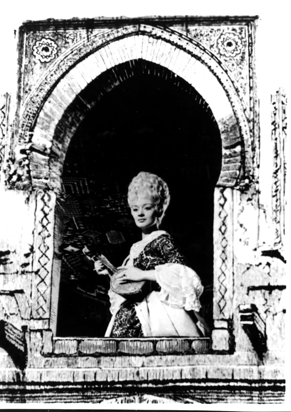 A black-and-white photo of a woman with an ornate hairdo holding an instrument in an arched window