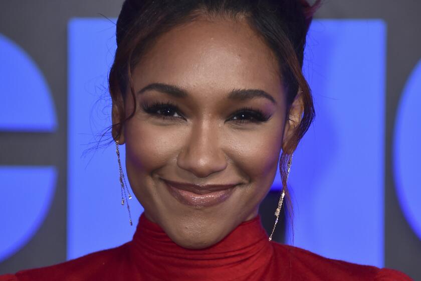 A woman wearing a red turtleneck smiles for cameras at a red carpet event. 