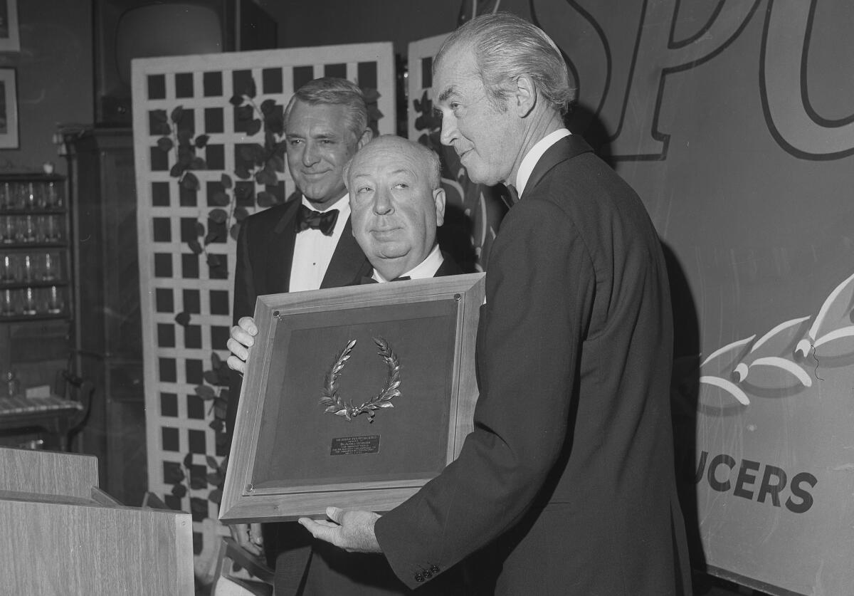 Alfred Hitchcock looks at Jimmy Stewart, who holds a large plaque. Cary Grant stands nearby.