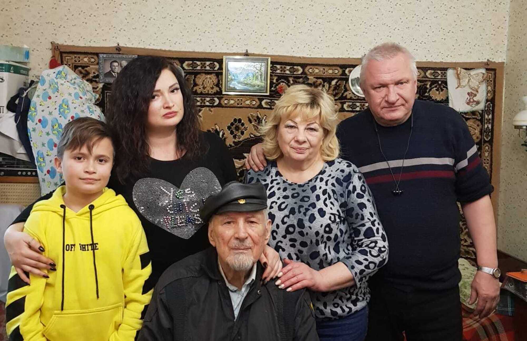 A family photo of an elderly man sitting, surrounded by a child, two women and a man, all standing.