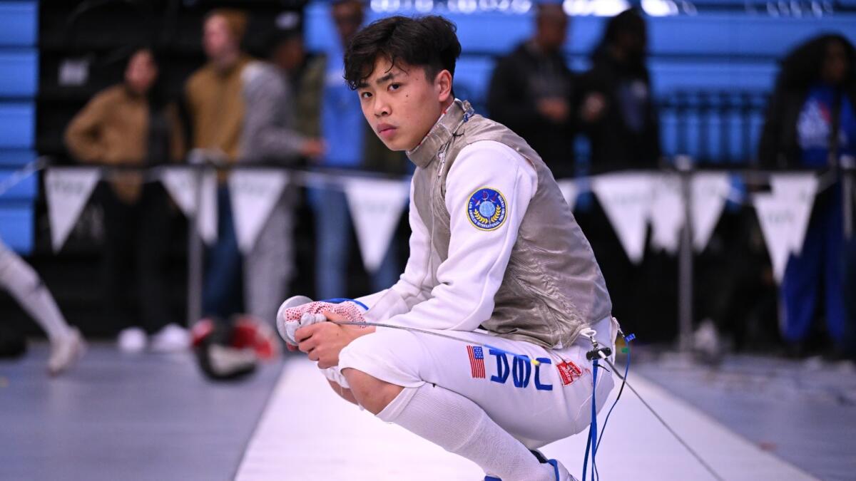 Bryce Louie squats on the floor in fencing garb, his foil lying across his knees