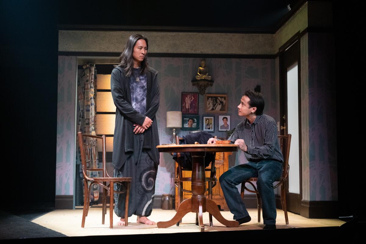 A man with long hair and bare feet stands beside a man seated at a small table in a living room setting on a stage.