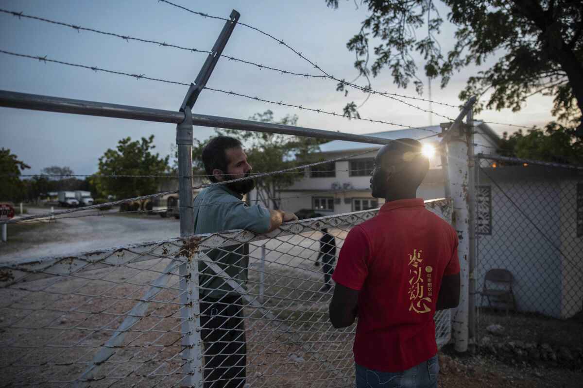 Two men speak from opposite sides of a chain link fence