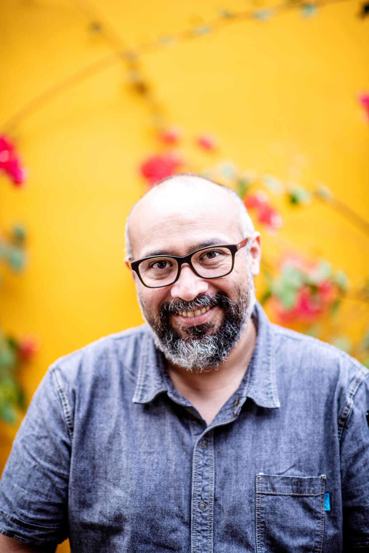 A portrait of Gilberto Cetina wearing glasses and a denim shirt against a yellow wall.
