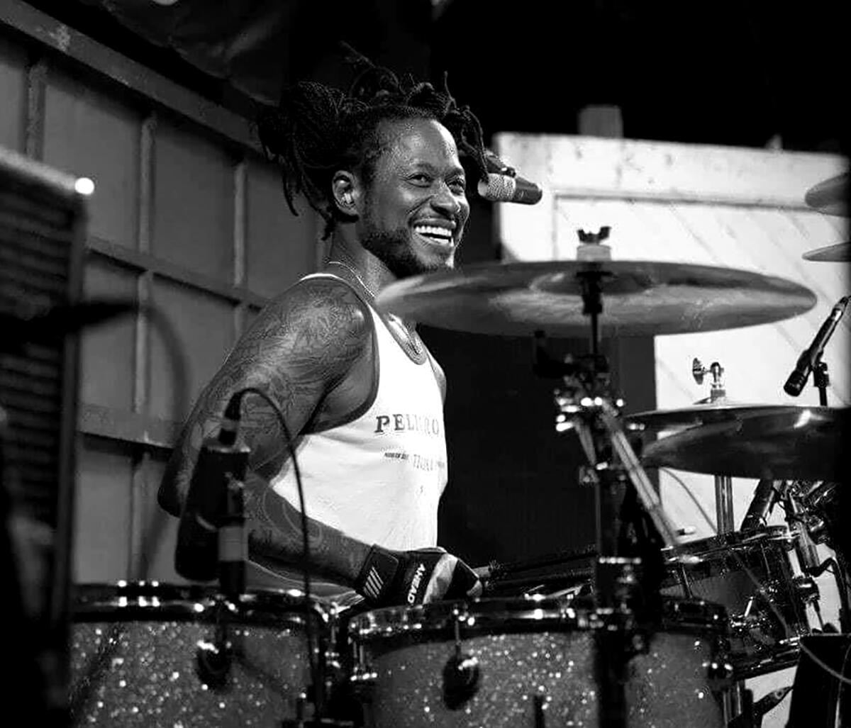 A black and white photo of D.H. Peligro in a white tank top smiling behind a drum kit.