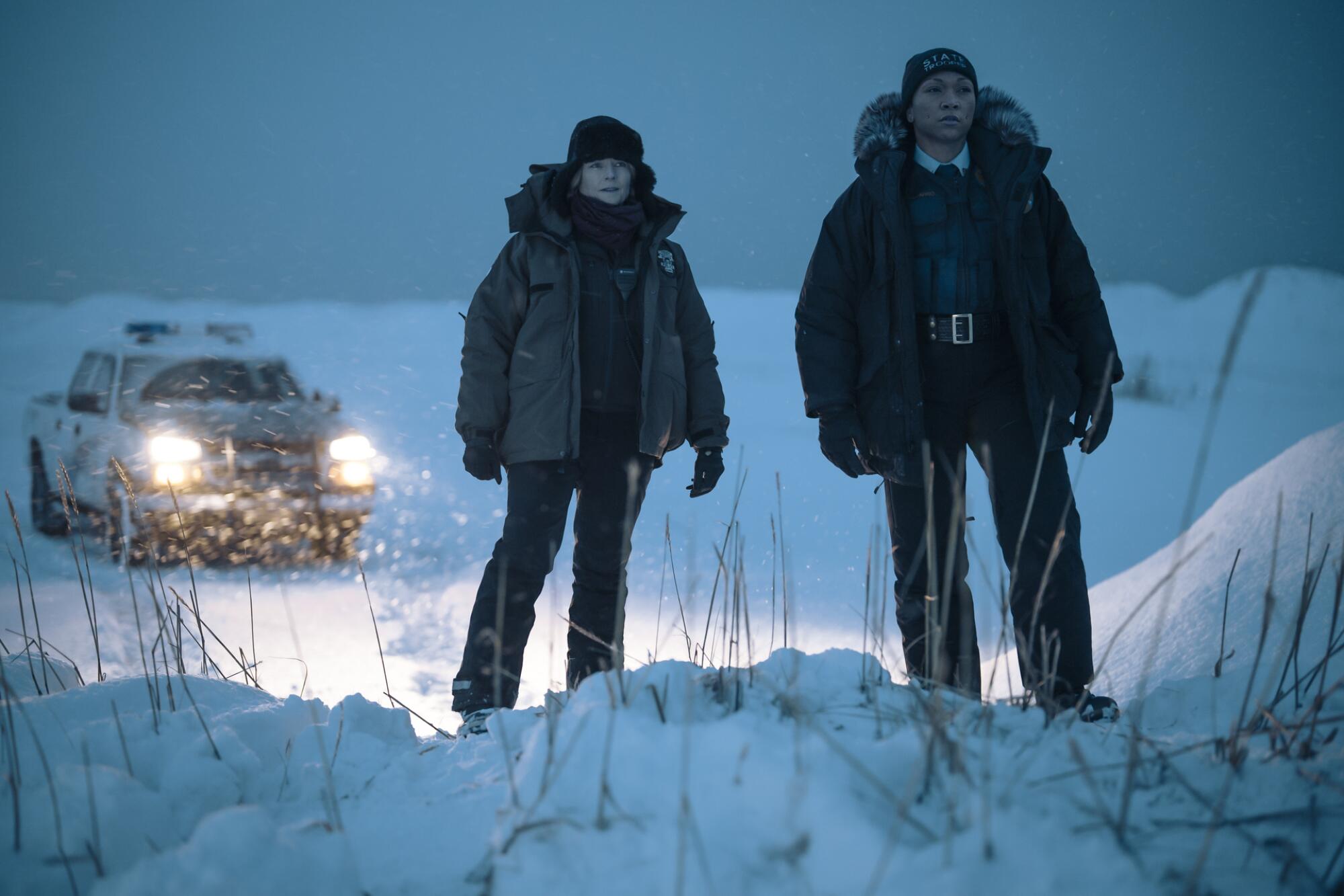 Two police officers at night, illuminated by the headlights of their truck, on a pile of snow.