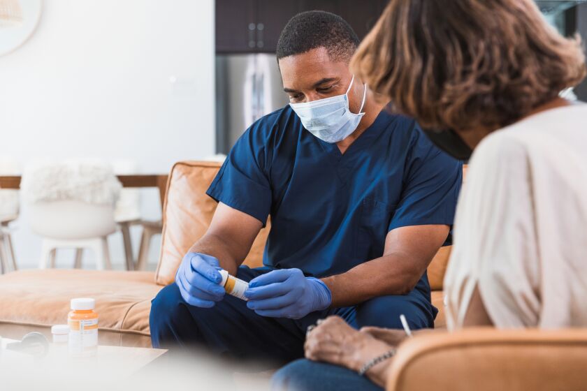 The mid adult male nurse wears protective gloves and mask as he sits in the woman's living room. The senior patient also wears a protective mask. They are being careful due to COVID-19.