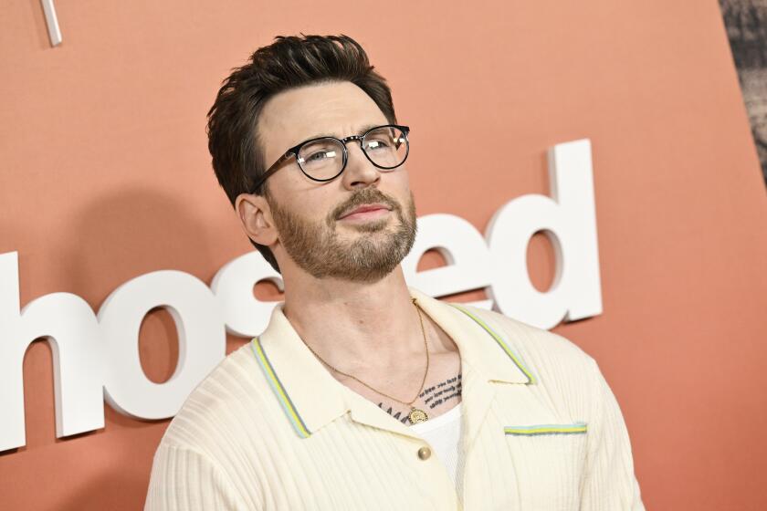 Chris Evans poses in glasses and a cream collared shirt against an orange background.