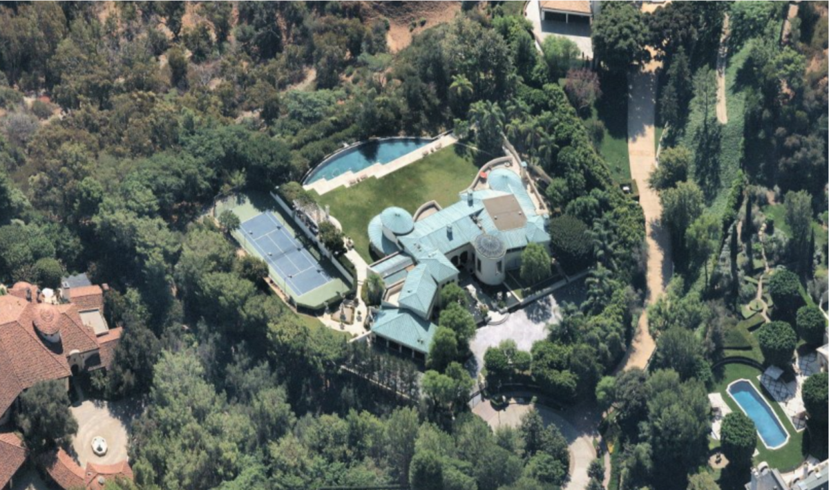 An aerial view of an estate shows a 15,300-square-foot mansion with tennis court, a large lawn and trees surrounding it all.