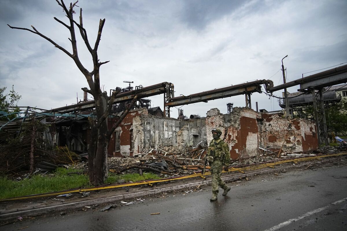 A Russian soldier walks along a road with a destroyed building behind him.