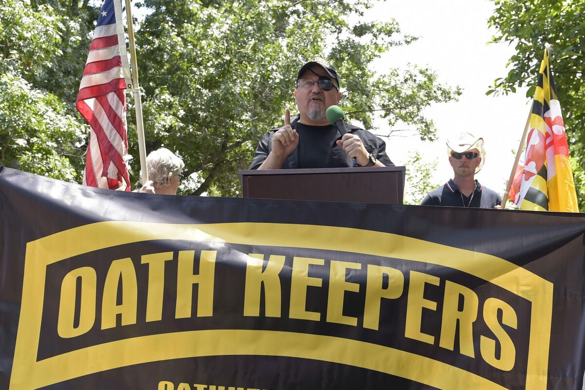 A man wearing an eyepatch speaks in front of an Oath Keepers banner