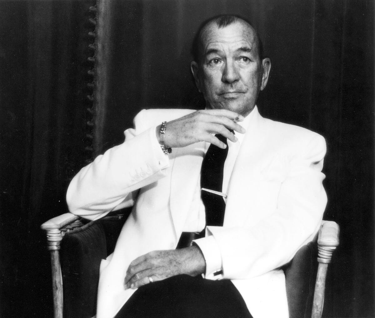 Noel Coward, wearing a white formal jacket, sits in a wooden chair.