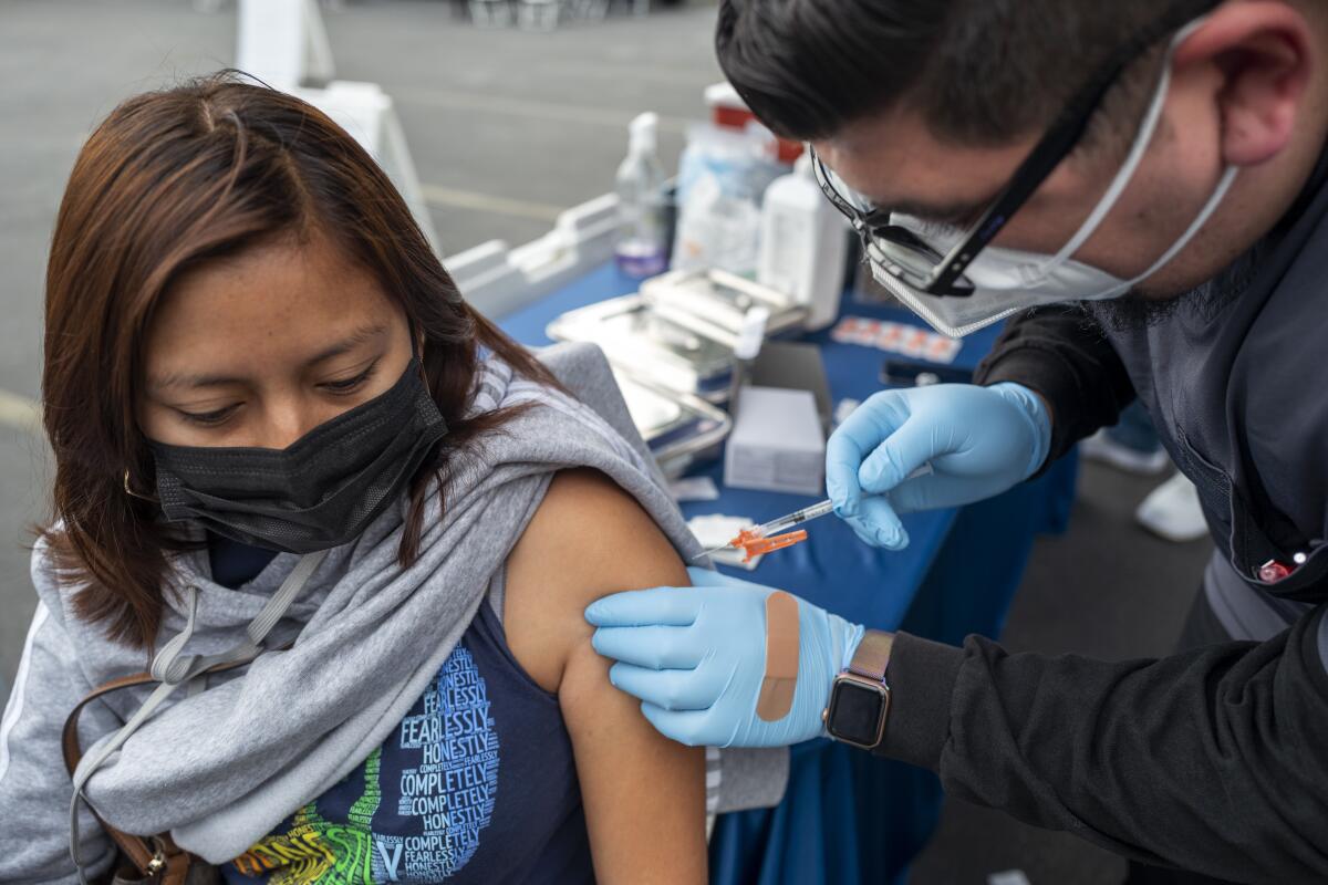 A man in glasses, plastic gloves and mask administers an inoculation to a woman in a mask.