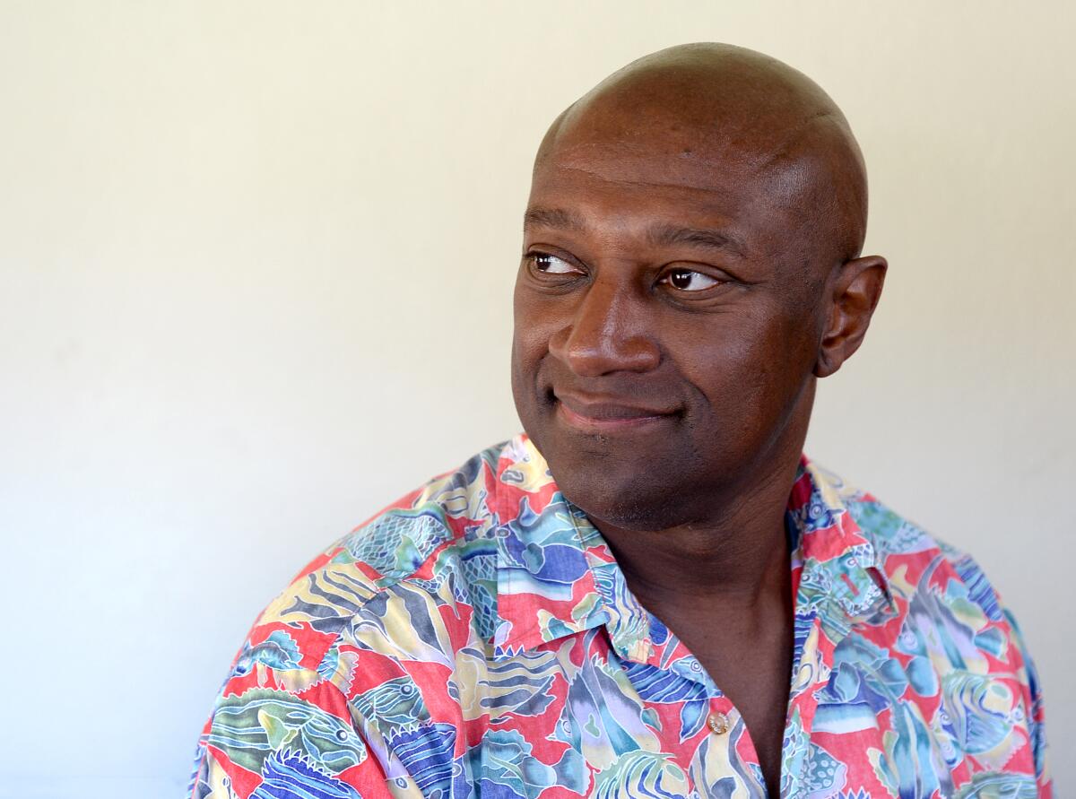 A man with a clean-shaven head, in a colorful pink, green and blue Hawaiian shirt, looks to the left