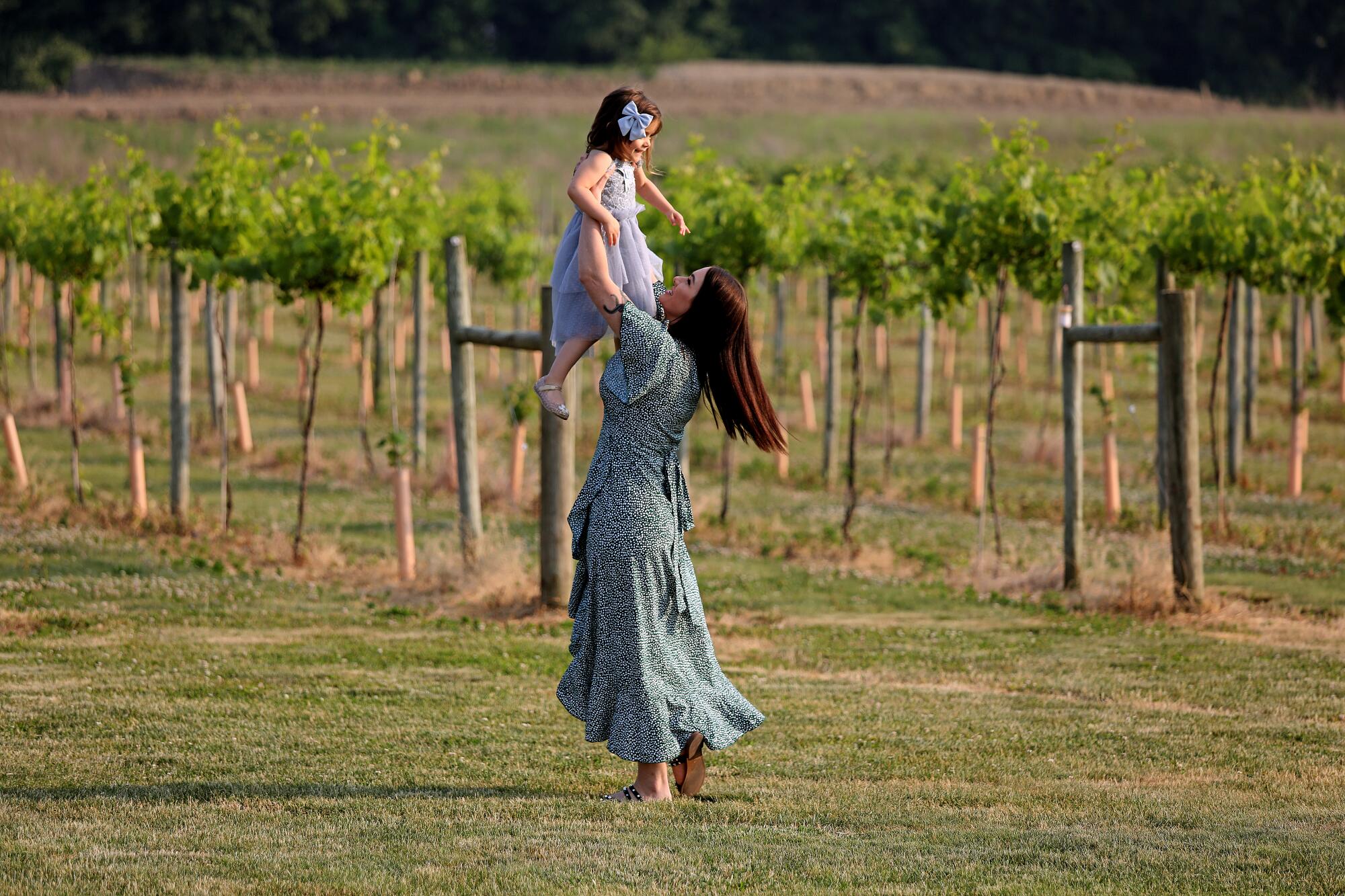 A woman holds a small child up in the air in a field next to a vineyard