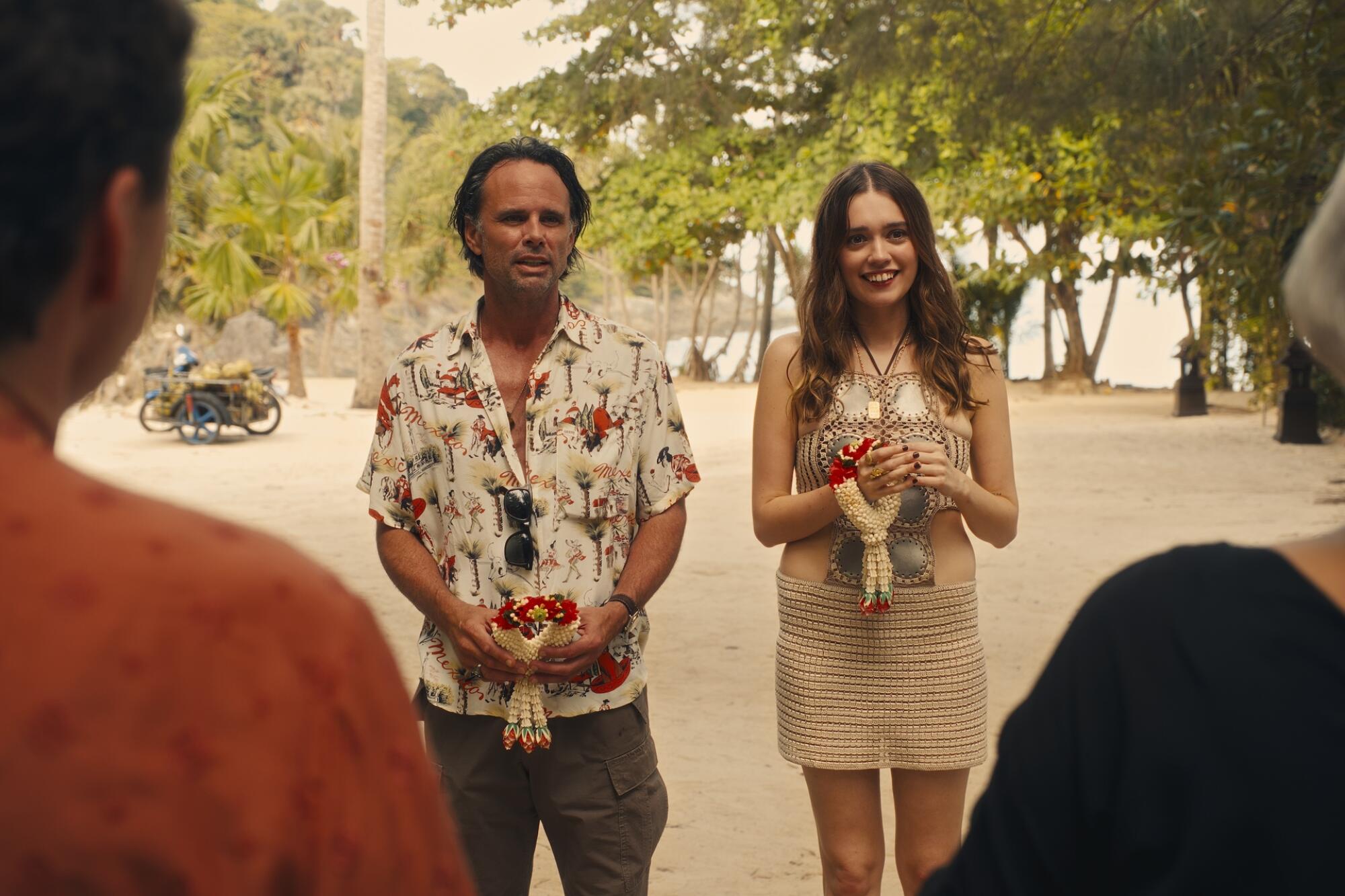 Walton Goggins, in a Hawaiian shirt and cargo shorts, stands next to Aimee Lou Wood, in a white halter top and skirt.
