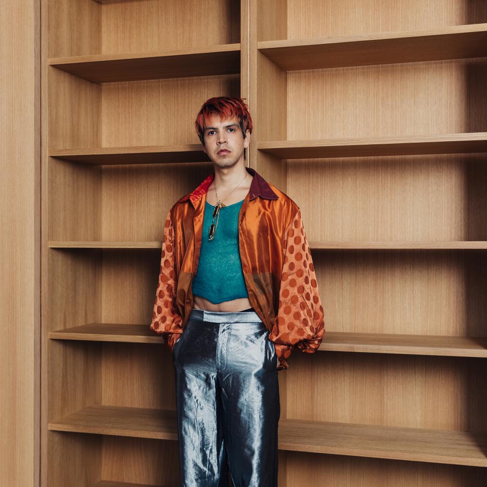 An person in shiny silver pants, green tank and orange shirt stands with hands in pockets in front of empty shelves.