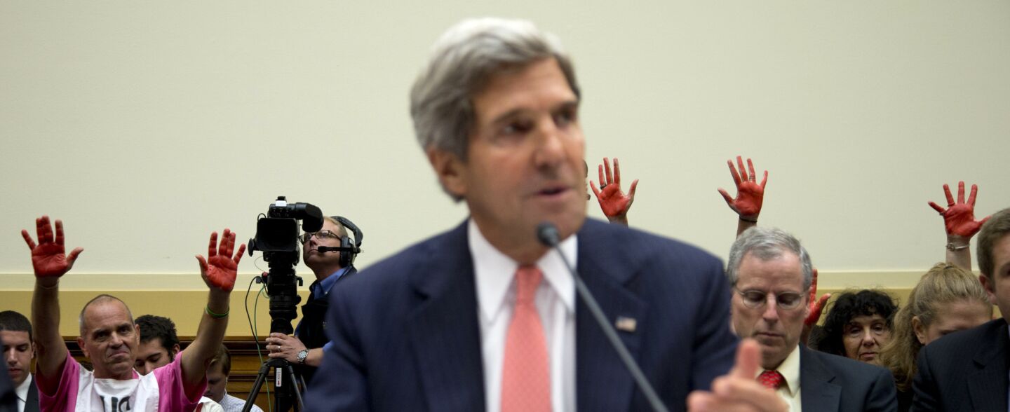 Protestors holding up their red-painted hands stand behind Secretary of State John Kerry as he testifies on Capitol Hill before a House Foreign Affairs committee hearing on U.S. military strikes on Syria.