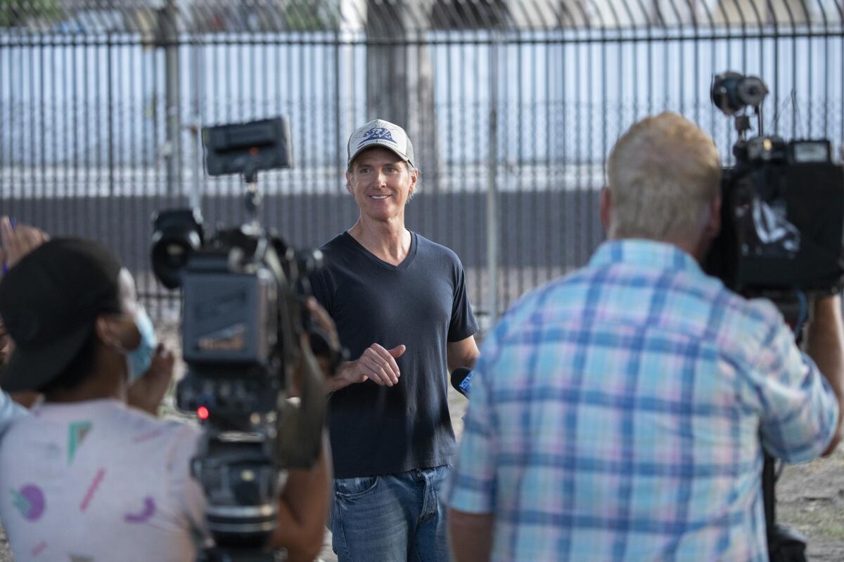 People pointing news cameras at Gov. Gavin Newsom as he stands near a tall security fence in a ball cap, T-shirt and jeans