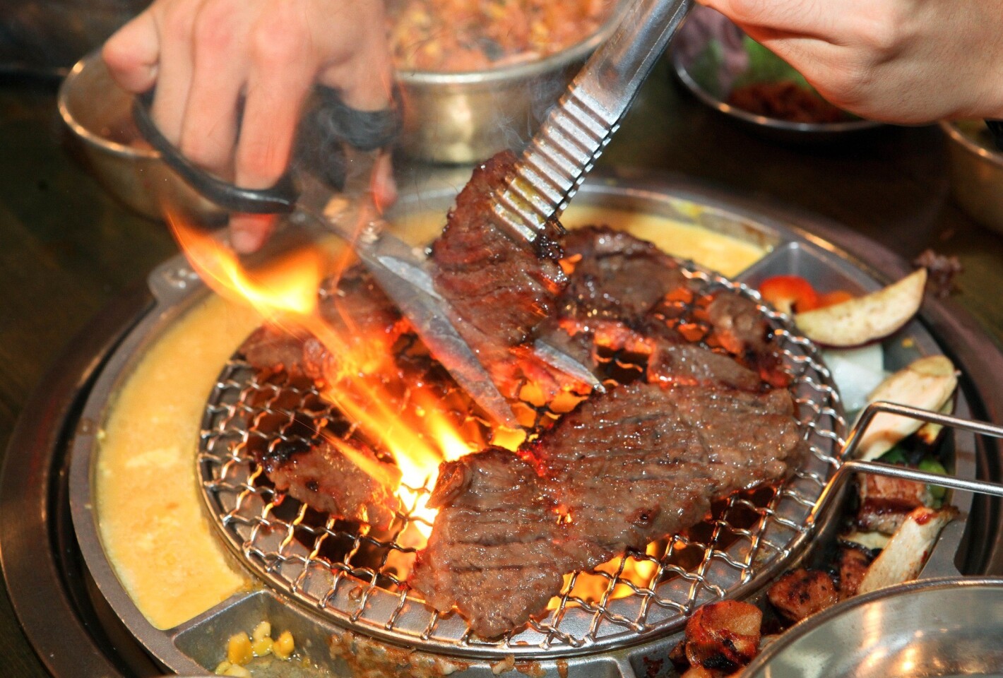 Portions of galbi beef are cut into smaller pieces as it cooks on a tabletop burner. Beaten egg and corn with cheese are also arrayed around the burners.