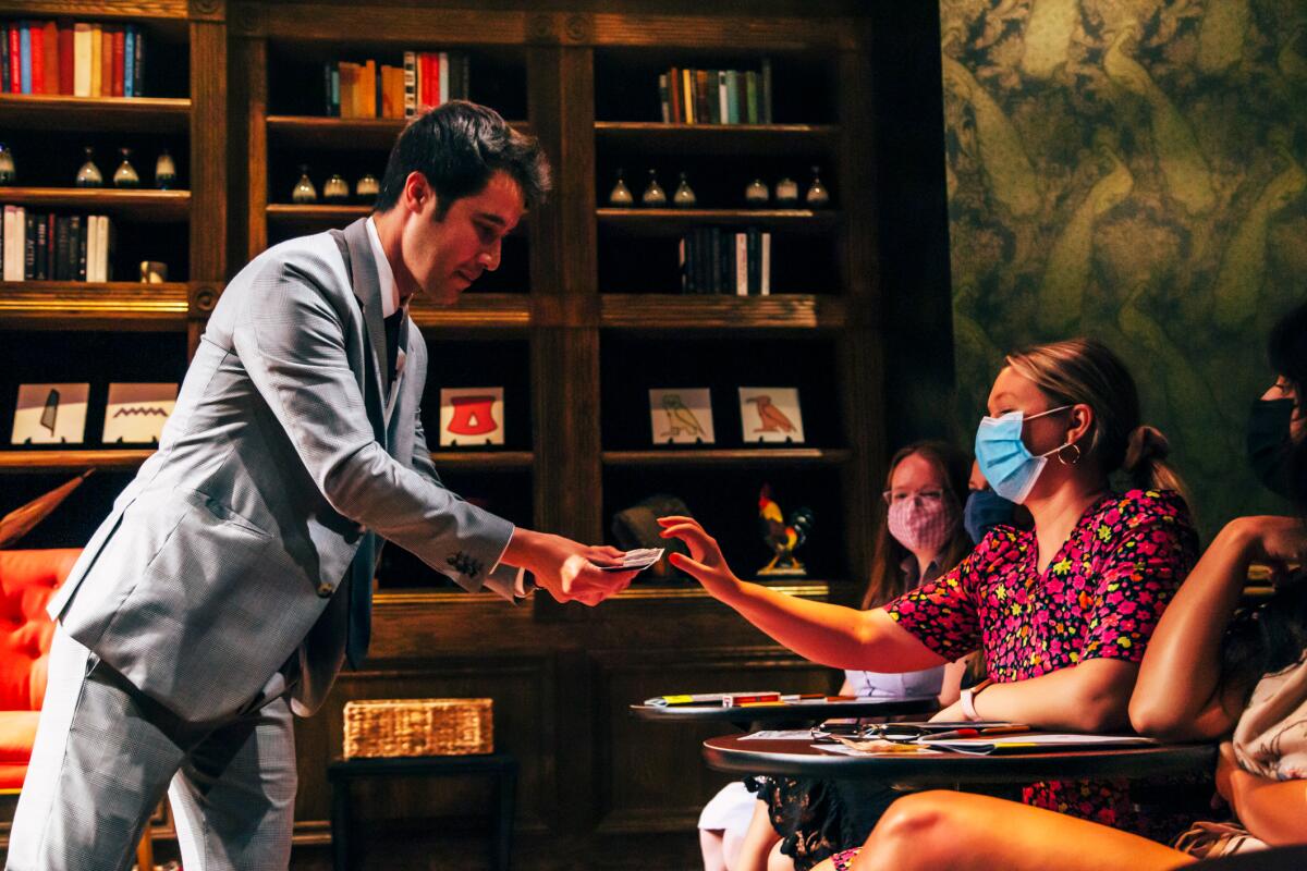 A magician extends a fan of playing cards to a masked audience member.