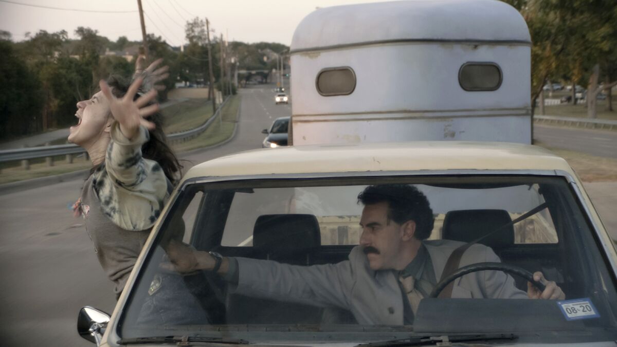 As Sacha Baron Cohen drives, Maria Bakalova leans out of a car window and screams.