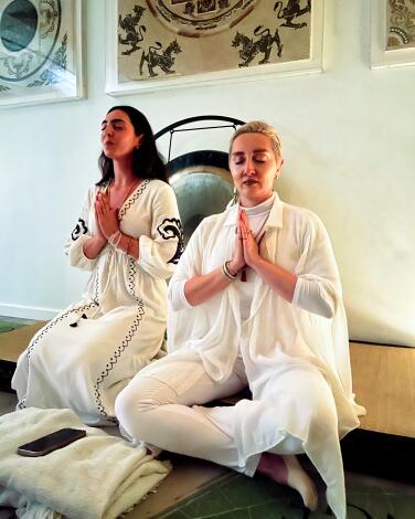Two people dressed in white hold their hands in a prayer position as they sit cross-legged on the floor next to a gong