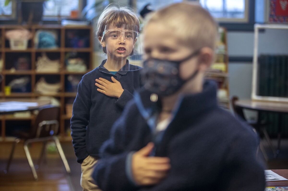Two children in anti-COVID masks stand with their hands over their hearts in a classroom.