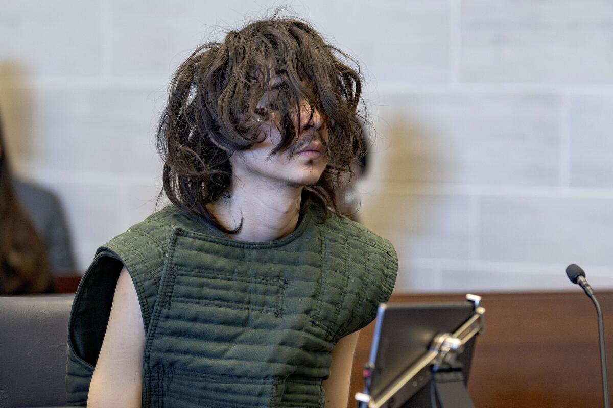 A slight young man sits in court wearing a green protective vest, his unkempt hair covering most of his face 