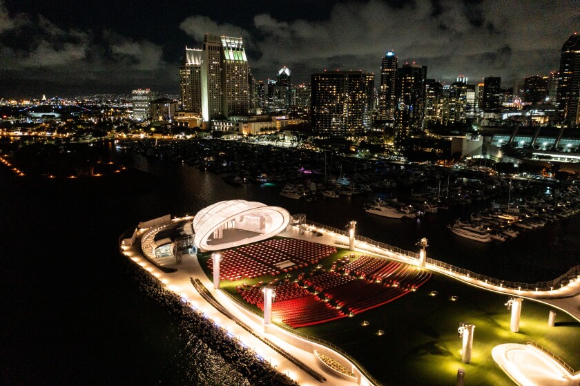 An illuminated white band shell is seen from overhead at night with a view of the San Diego skyline in the distance.