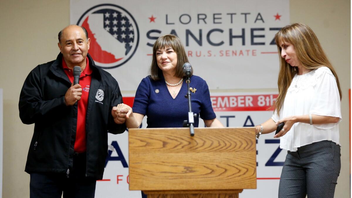 A dark-haired woman in a navy blue suit stands at a lectern, flanked by a man holding a microphone and a woman in a white blouse.