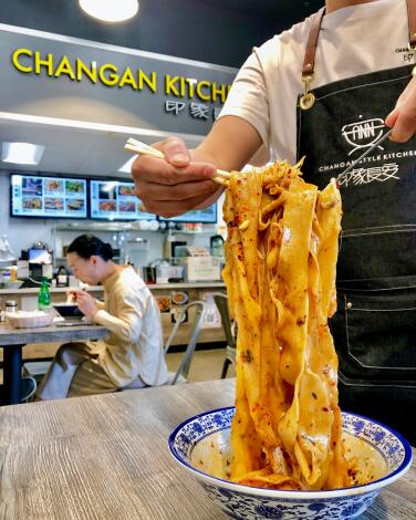 A customer eats at a food court table as a person wearing an apron lifts wide noodles in a red sauce from a bowl using chopsticks