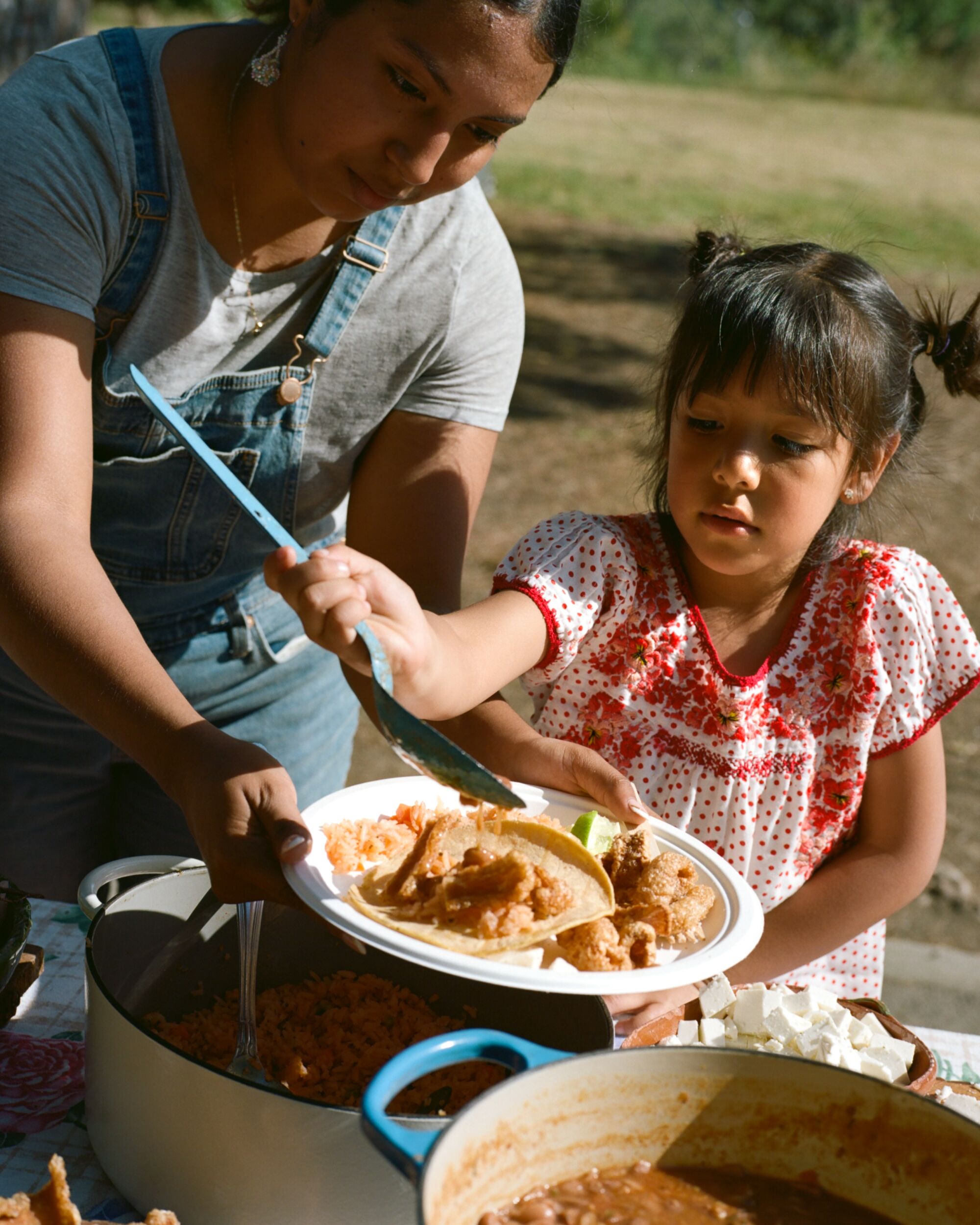 A woman helps a young girl put food on a plate in a park.