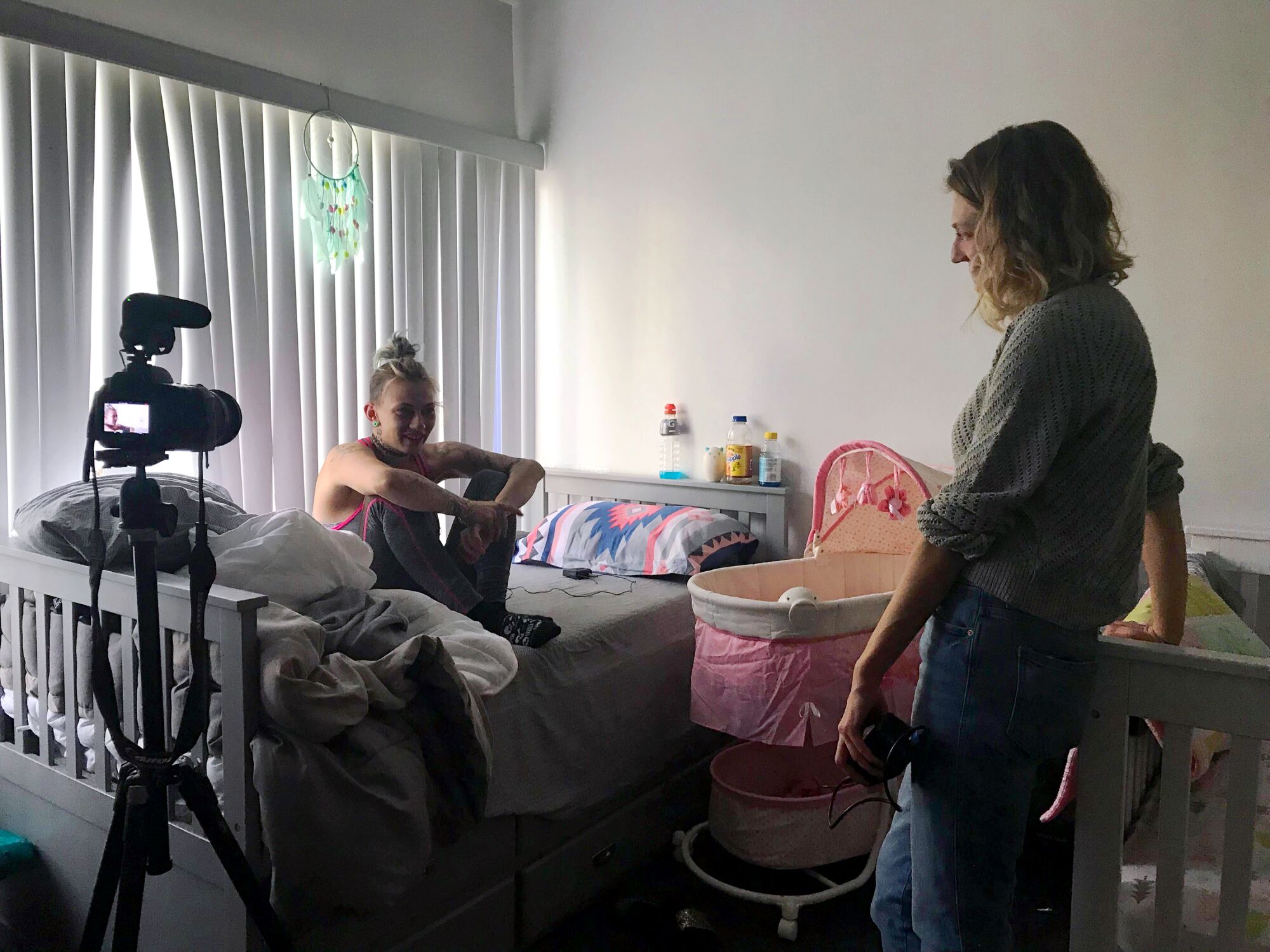 A woman seated on a bed with a camera pointed at her, while another woman stands smiling at her.