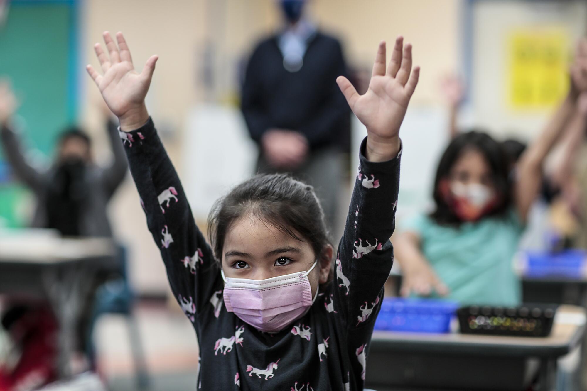 A girl in a pink face mask lifts both arms into the air.