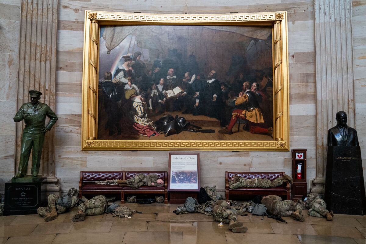In front of benches and a large historic painting, men in uniform sleep on the floor.