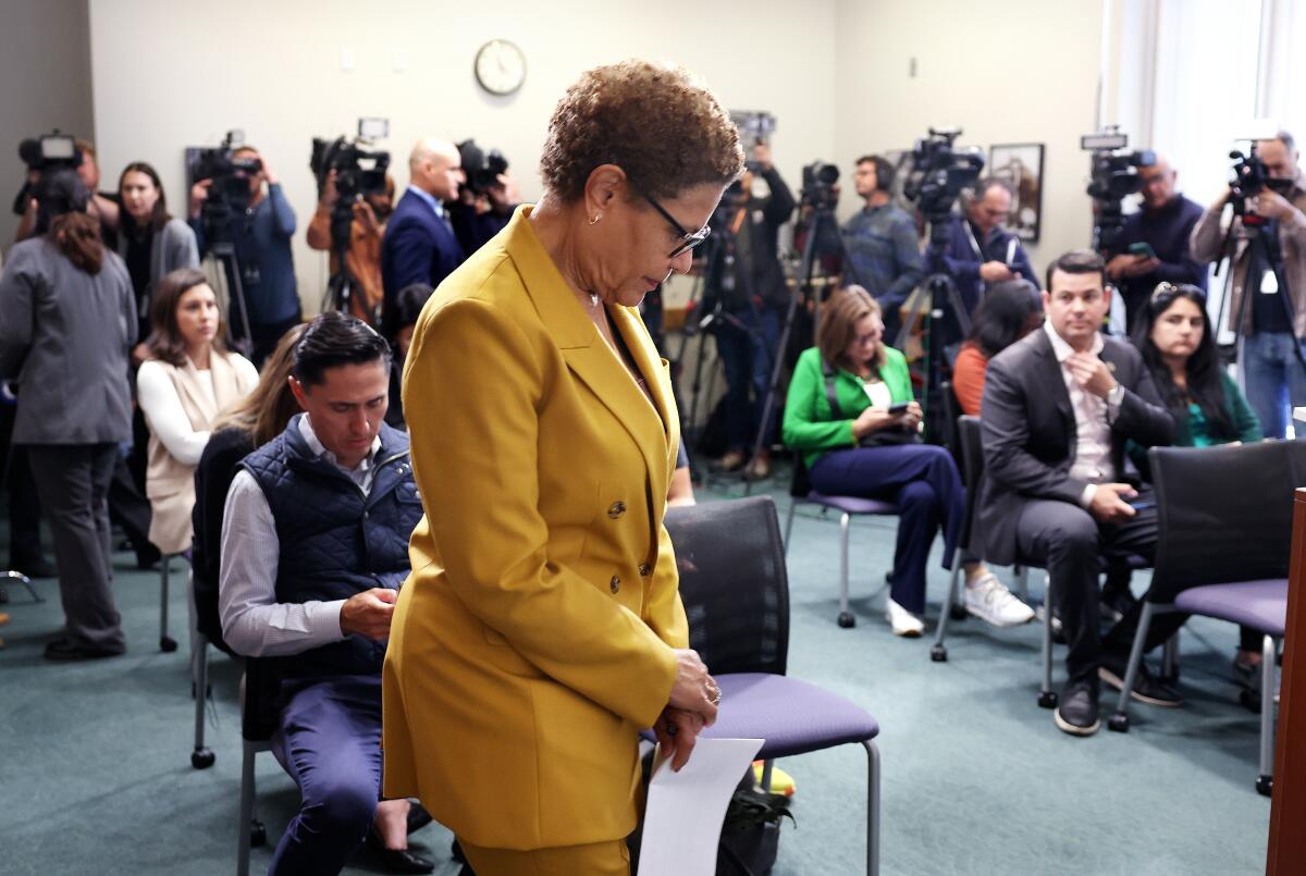 A woman with a bowed head in a room full of seated people and TV cameras in the back.