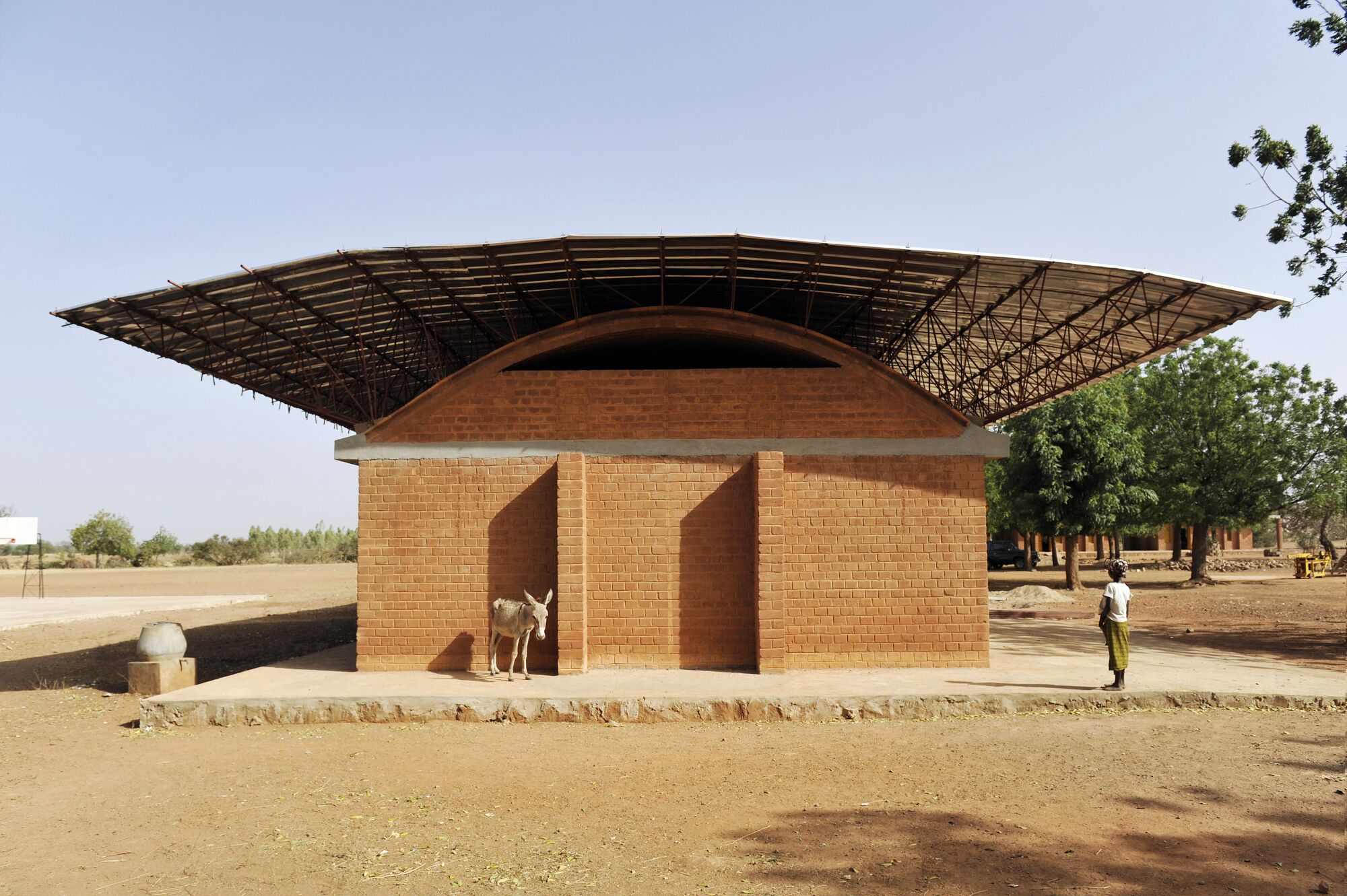 A donkey and a woman stand before a one-story clay brick building that is sheltered by a floating roof.