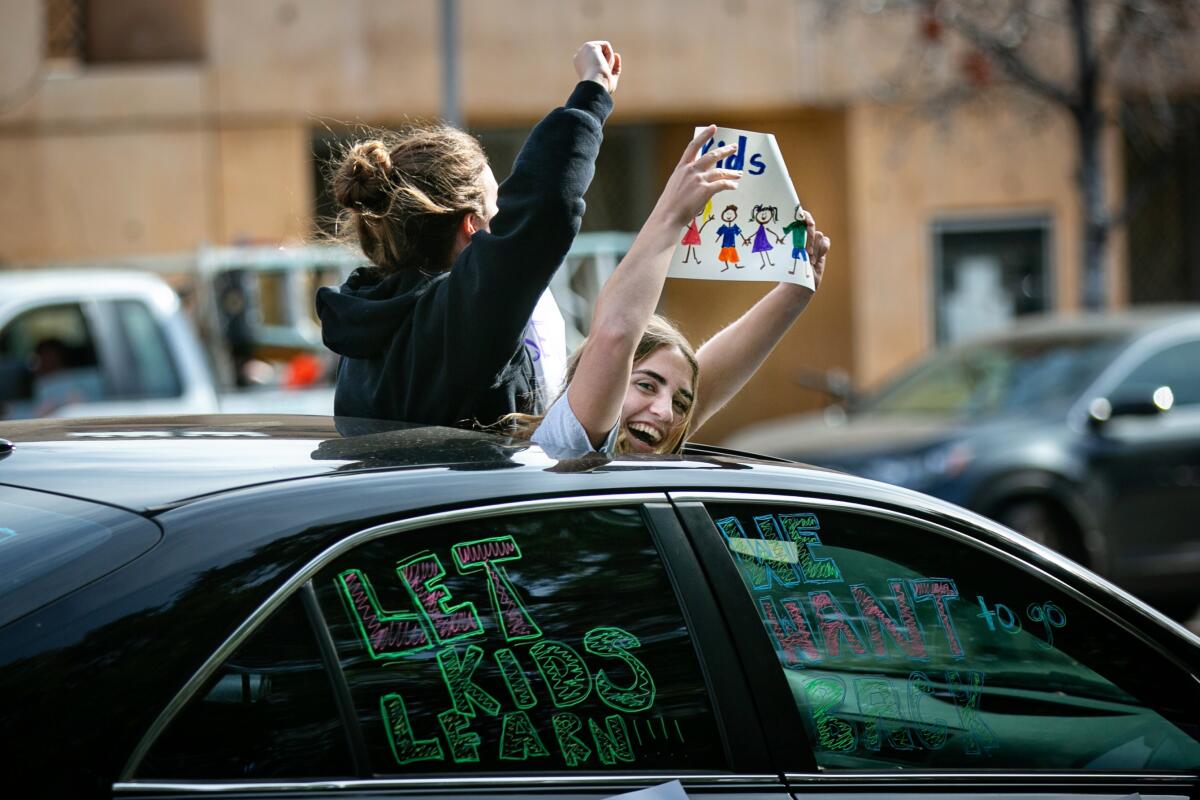 Two girls standing out of a car's sunroof hold signs. The car's windows read Let kids learn, we want to go back