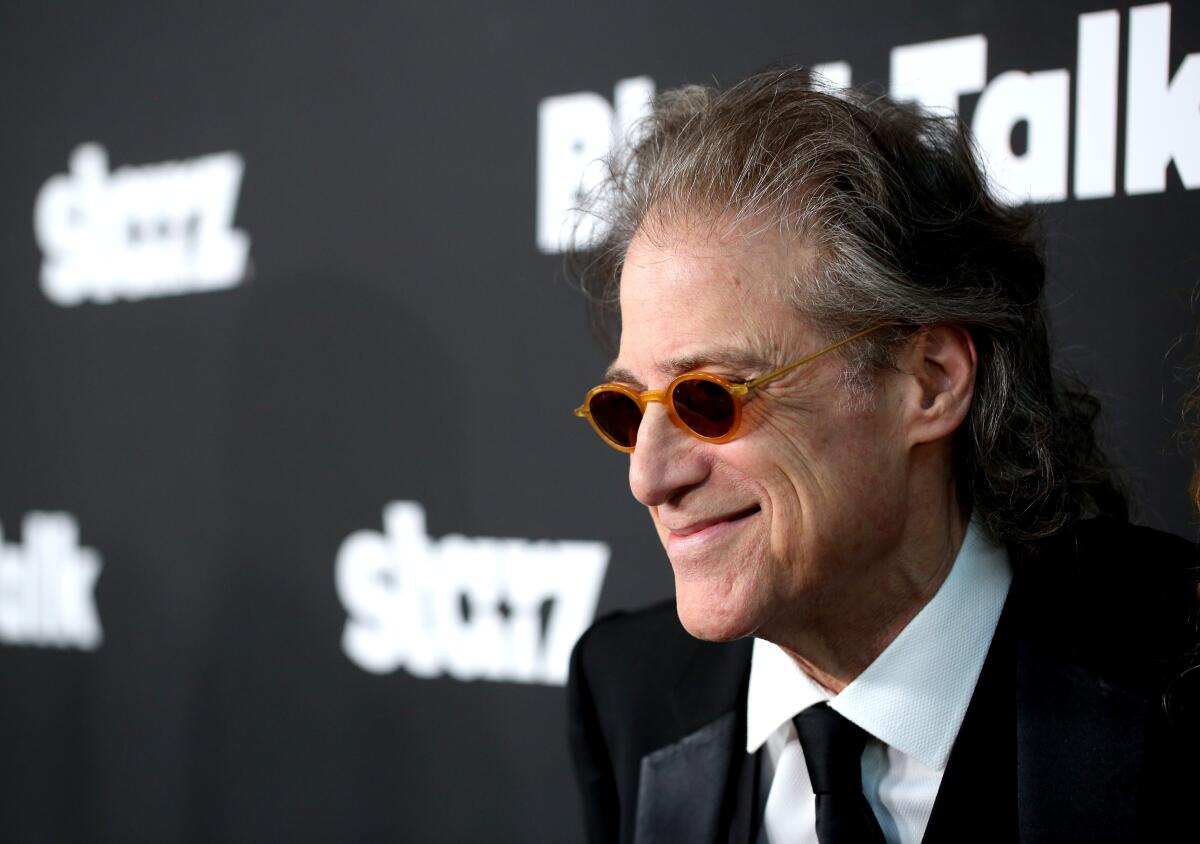 Richard Lewis wears yellow-rimmed sunglasses and smiles in a suit while standing front of a backdrop