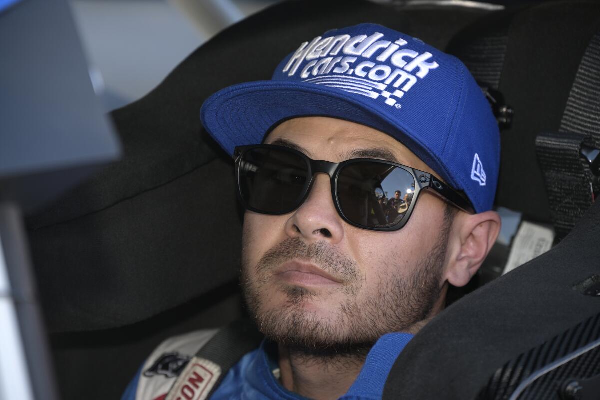 Driver Kyle Larson sits wearing sunglasses and a baseball cap in his car on pit road before a NASCAR race.