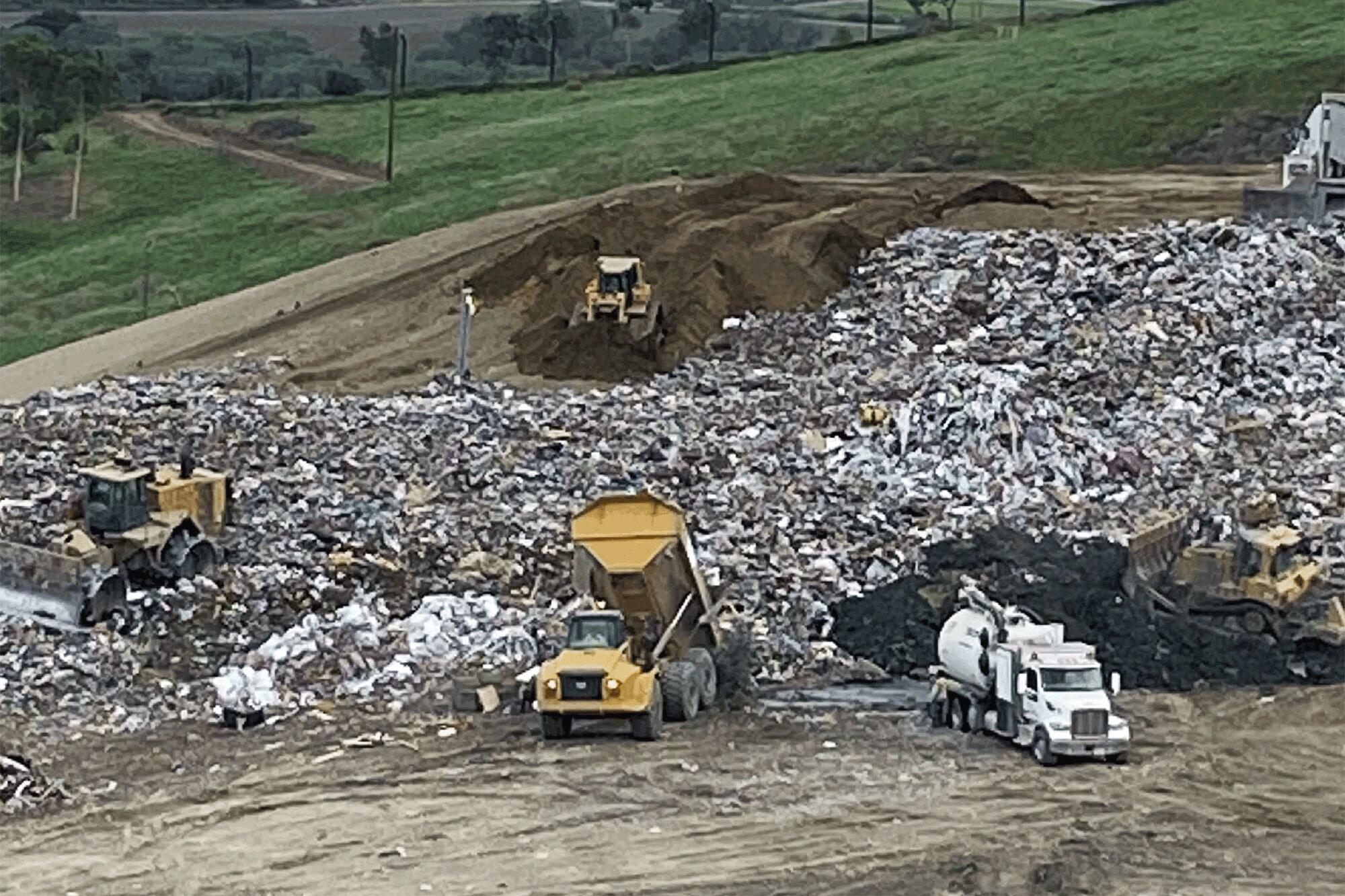 Bulldozers and dump trucks move piles of trash and dirt at a landfill.