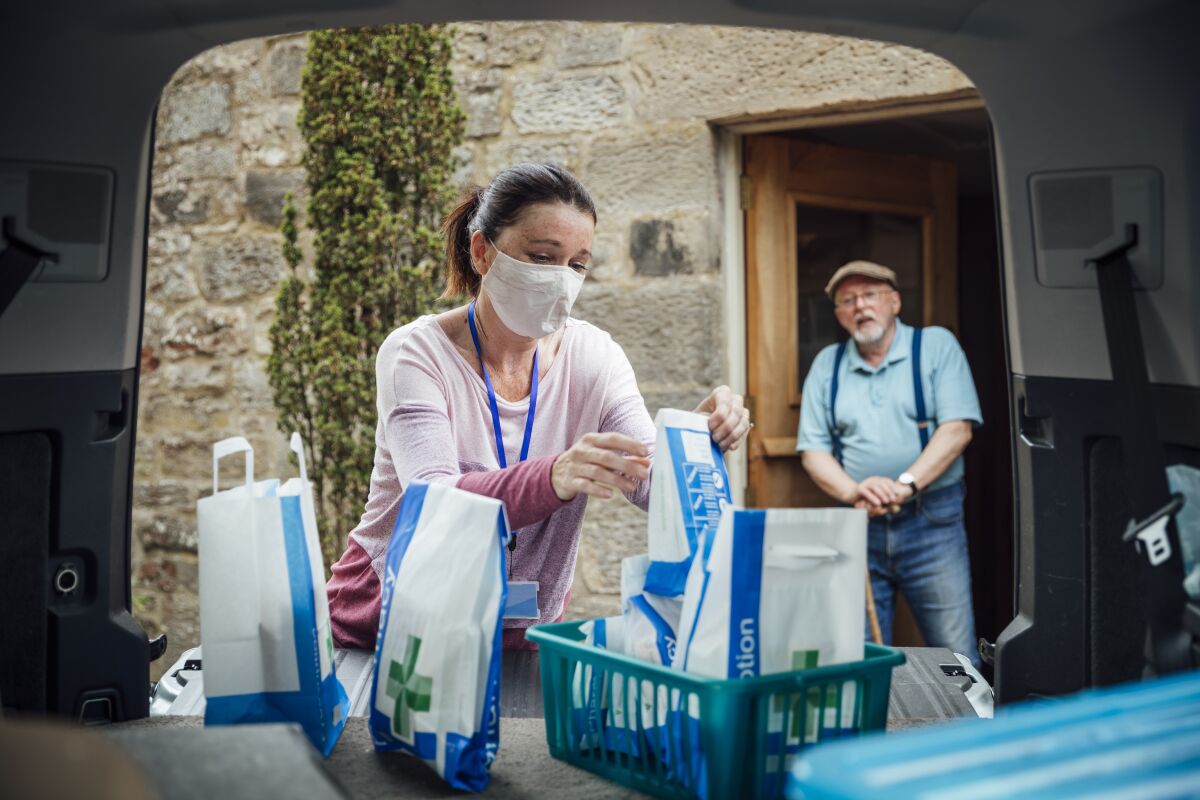 A woman wearing a mask brings supplies and prescriptions from the pharmacy to an older man at his home.