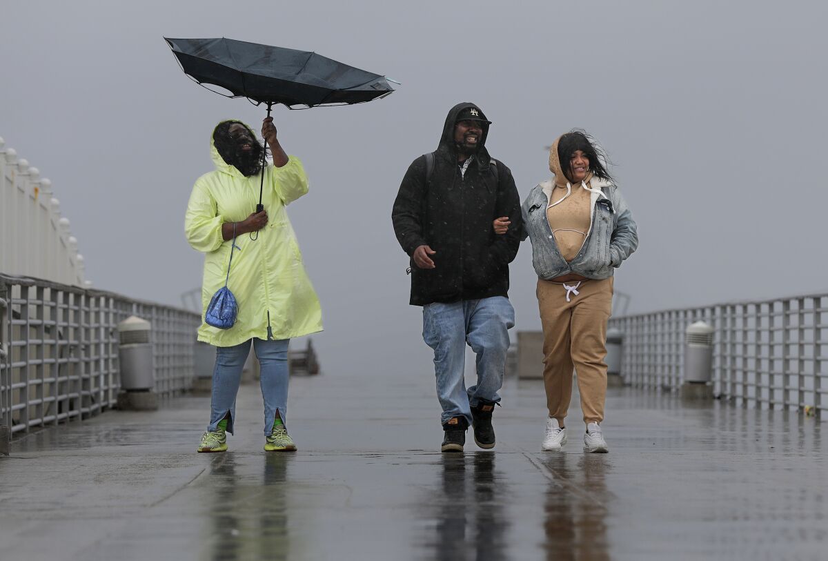A woman holds an umbrella overturned by the wind while walking with two other people on a wet pier