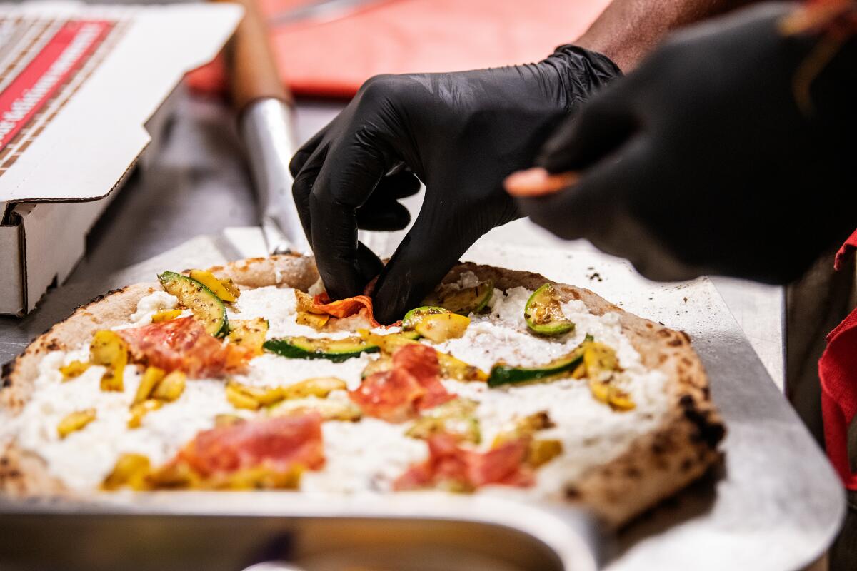 Hands in black rubber gloves place toppings on a pizza.