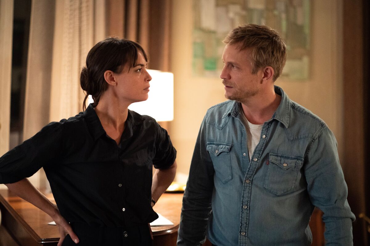A woman and a man talking in a room with a lamp on a table behind them.
